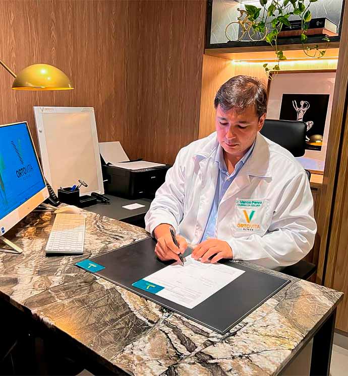 Dr. Marcio Penna de Carvalho, ortopedista especialista em coluna, escrevendo em um papel, sentado em sua mesa.